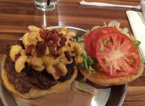 A burger with a beef patty topped with macaroni and cheese, bacon, and a side of fresh lettuce and tomato on a bun.