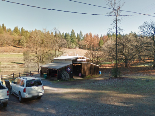 A rustic wooden building surrounded by trees and a gravel driveway, with a few parked vehicles nearby.