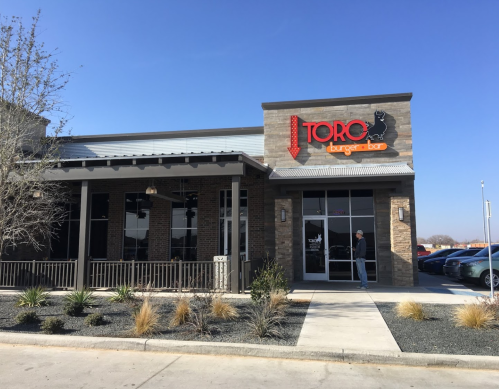 Exterior of Toro Burger Bar, featuring a modern design with a sign and outdoor seating area under a clear blue sky.
