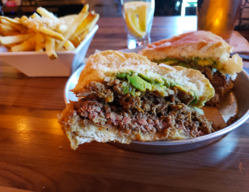 A close-up of a sandwich filled with shredded meat and avocado, served with a side of fries and a drink in the background.