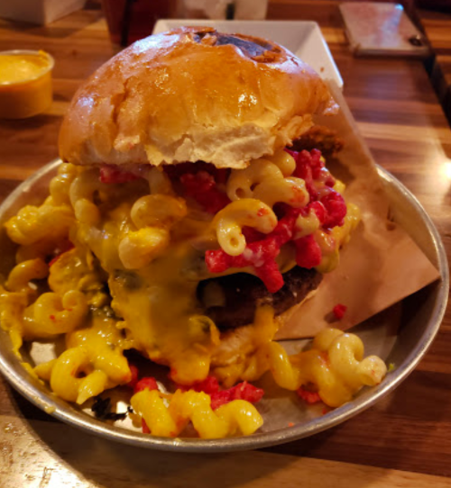 A towering burger topped with macaroni and cheese, Cheetos, and melted cheese, served on a metal plate.