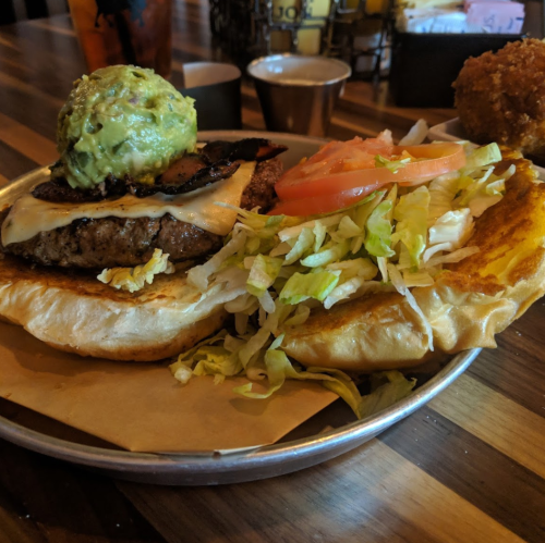 A close-up of a burger with melted cheese, guacamole, lettuce, and tomato on a toasted bun, served with a side.