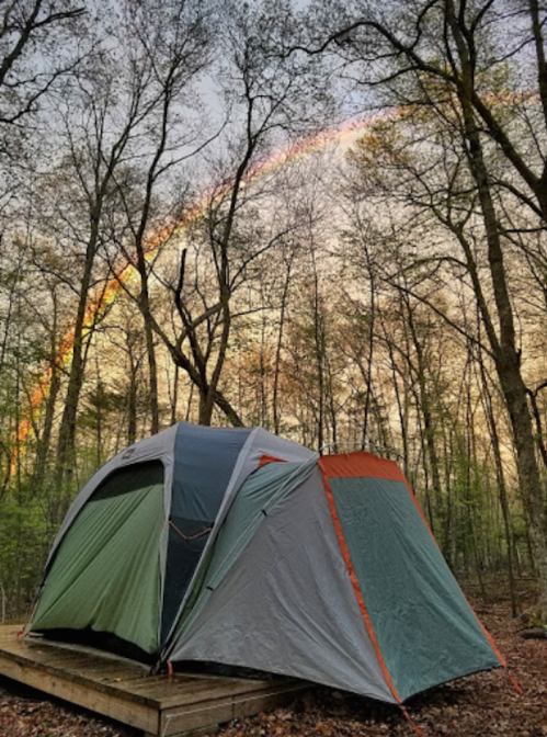 A tent on a wooden platform surrounded by trees, with a rainbow arching across the sky at sunset.