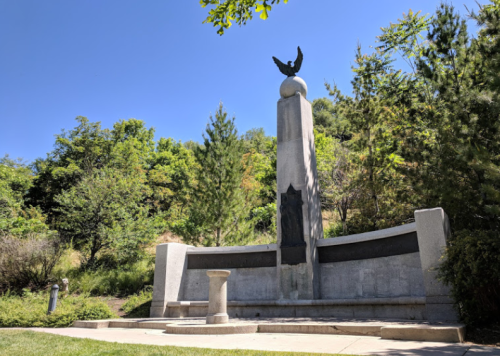 A stone monument with an eagle atop, surrounded by trees and greenery under a clear blue sky.