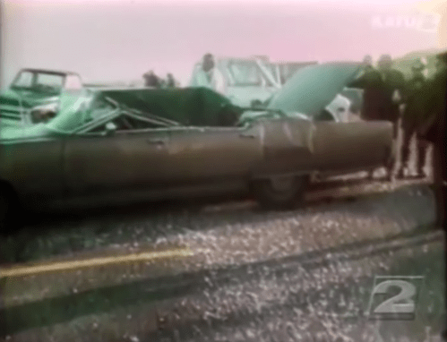 A damaged car with a crumpled roof, surrounded by onlookers at an accident scene.