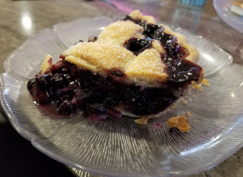 A slice of berry pie on a glass plate, with a golden crust and dark berry filling oozing out.
