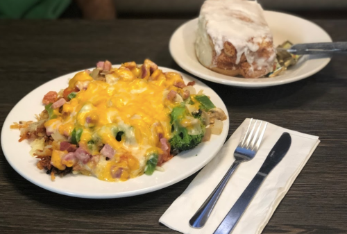 A plate of cheesy vegetable and ham hash next to a cinnamon roll with icing on a wooden table.