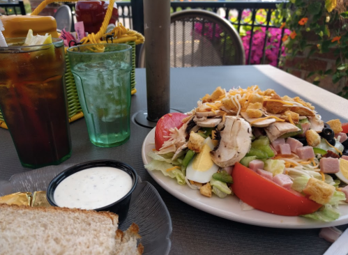 A colorful salad with chicken, ham, cheese, and croutons, served with drinks and a side of ranch dressing.