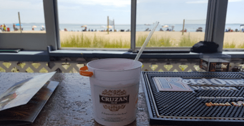 A beach view with a drink on a table, featuring sand, grass, and people in the distance under a clear sky.