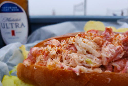 A close-up of a lobster roll topped with seasoning, served with a lemon slice and a bottle of beer in the background.