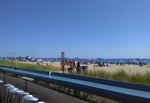 A sunny beach scene with people under umbrellas, sand, and a clear blue sky, viewed from a nearby deck.