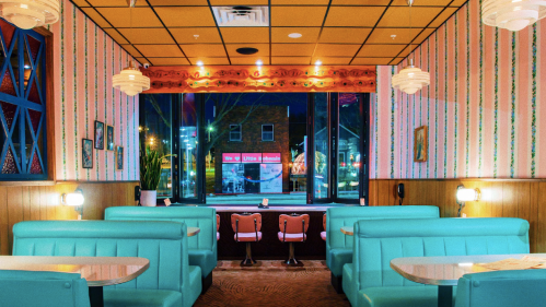 Retro diner interior with teal booths, warm lighting, and large windows showcasing a nighttime city view.