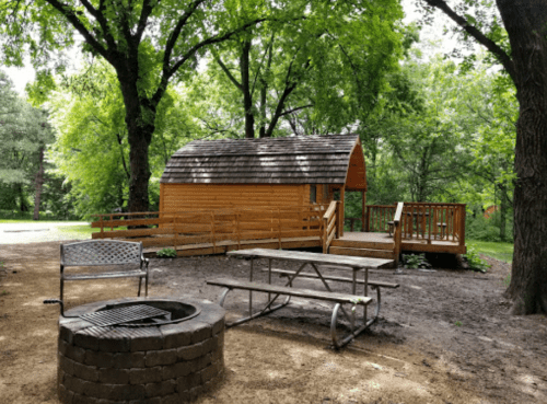 A wooden cabin surrounded by trees, with a picnic table, fire pit, and a small deck in a natural setting.