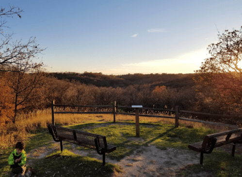 A scenic overlook with benches, surrounded by autumn trees and a child playing nearby at sunset.