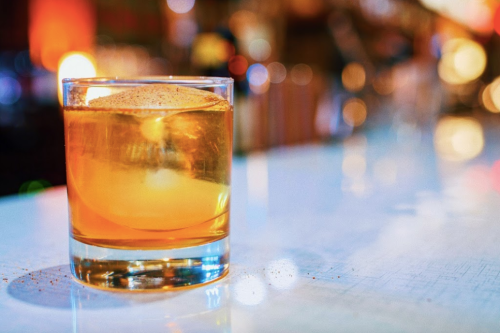 A glass of amber-colored whiskey on a bar counter, with a large ice cube and a sprinkle of cinnamon on top.