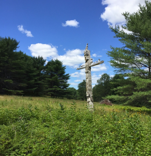 A weathered wooden sculpture stands in a grassy field, surrounded by trees and a bright blue sky with fluffy clouds.