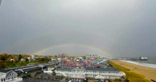 A coastal view featuring a roller coaster, beach, and a double rainbow under a cloudy sky.