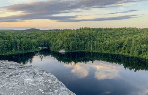 A serene landscape featuring a calm lake surrounded by lush green trees under a colorful sky at sunset.