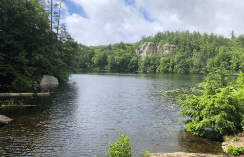 A serene lake surrounded by lush green trees and rocky cliffs under a partly cloudy sky.
