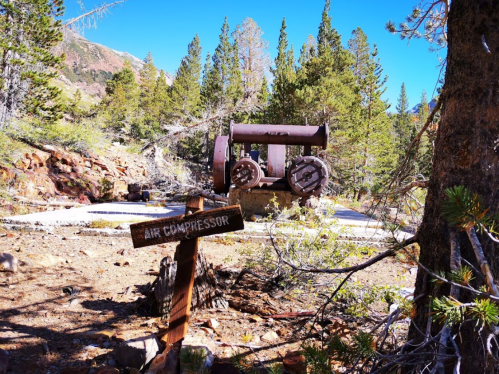 An old air compressor sits among trees in a rocky area, with a wooden sign pointing to it.