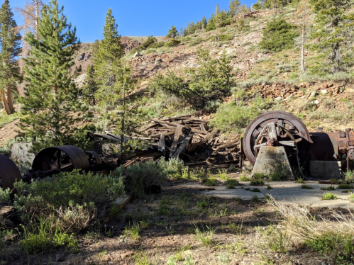 Abandoned machinery and wooden debris in a forested area with rocky terrain and trees in the background.