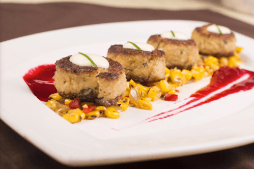 A plate of golden corn with four round crab cakes topped with sauce and garnished with chives, drizzled with berry sauce.