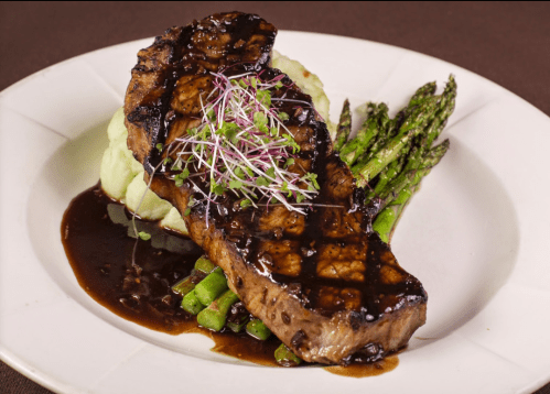 Grilled steak with a rich sauce, served on a bed of mashed potatoes and asparagus, garnished with microgreens.