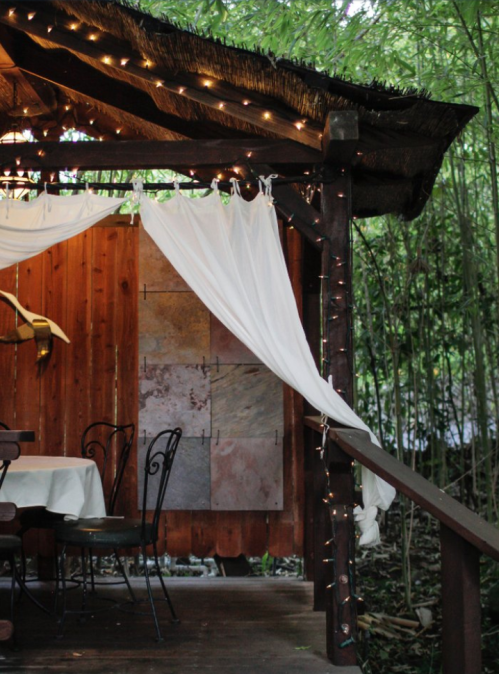 A cozy outdoor space with a wooden structure, draped white fabric, and string lights surrounded by bamboo.