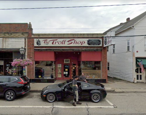 A storefront of "The Troll Shop" with two people outside and parked cars on the street. Flowers hang from the building.