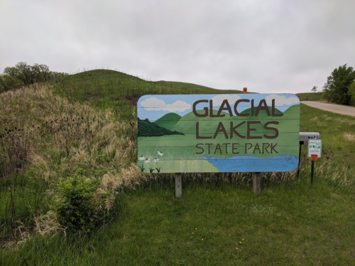Sign for Glacial Lakes State Park, featuring a painted landscape with hills and greenery, near a gravel road.