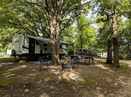 A camper parked under trees with a picnic table and fire pit nearby, surrounded by a natural, wooded area.