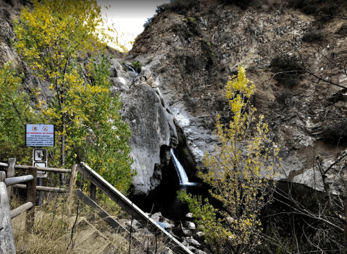 A scenic view of a waterfall surrounded by rocky cliffs and autumn foliage, with a warning sign nearby.
