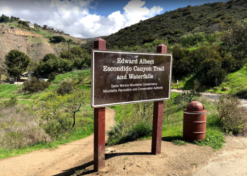 Sign for Edward Albert Escondido Canyon Trail and Waterfalls, surrounded by greenery and hills under a cloudy sky.