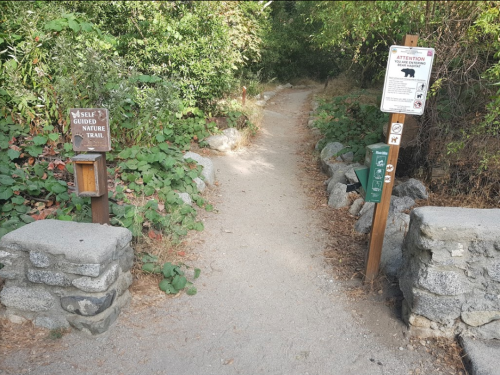 A dirt path leads into a wooded area, flanked by signs for a self-guided nature trail and wildlife warnings.