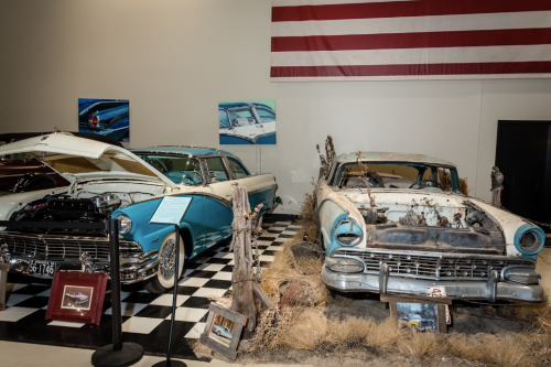 Two vintage cars in a museum: a restored turquoise model and a weathered, rusty one, set against an American flag backdrop.