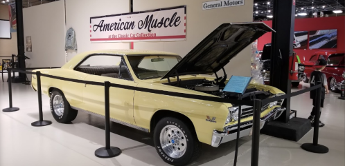 A classic yellow muscle car on display with its hood open, surrounded by barriers in a car collection exhibit.