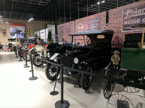A row of vintage cars displayed in a museum, with a brick wall backdrop and historical signage.