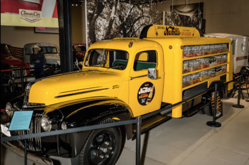 A vintage yellow delivery truck with a soda display on the side, showcased in a museum setting.