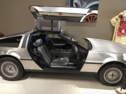 A silver DeLorean car with gullwing doors open, displayed in a museum setting.