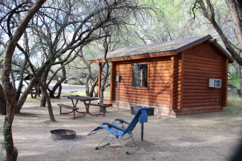 A rustic log cabin surrounded by trees, with a picnic table, a campfire ring, and a blue chair nearby.