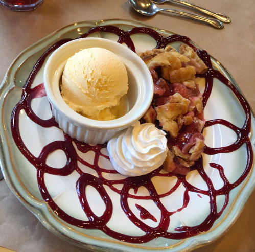 A dessert plate featuring a slice of pie with berry sauce, a scoop of vanilla ice cream, and whipped cream.