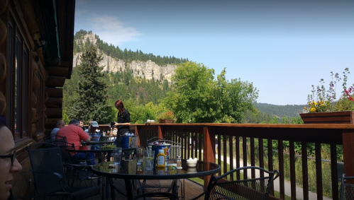 A scenic outdoor dining area with patrons, overlooking a mountainous landscape and greenery.