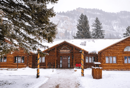 A cozy log cabin restaurant surrounded by snow-covered trees and mountains on a winter day.