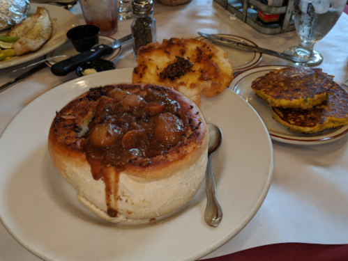 A bowl of stew served in a bread bowl, with a side of cornbread and pancakes on a white plate.