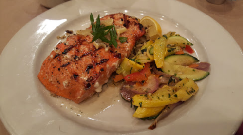 Grilled salmon fillet garnished with herbs, served with a side of sautéed vegetables and lemon slices.