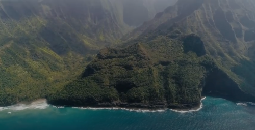 Aerial view of lush green mountains meeting a blue ocean along a rugged coastline.