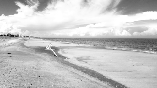 A black and white beach scene with a tilted sign, gentle waves, and clouds in the sky.