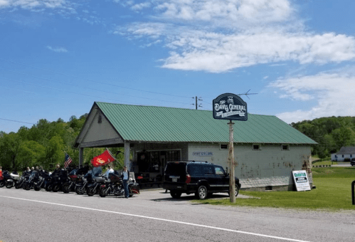 A small general store with a green roof, surrounded by parked motorcycles and flags, under a blue sky.