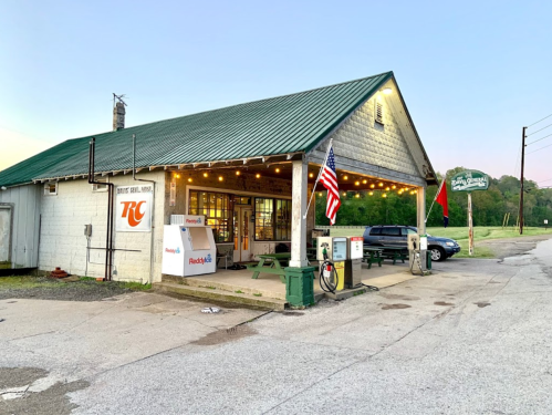 A rustic gas station with a green roof, string lights, and an American flag, set against a twilight sky.