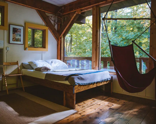 Cozy bedroom with a wooden bed, hammock, and large windows overlooking a forested area. Warm, inviting atmosphere.